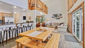 Een ruime woonkamer met een houten eettafel, hoog plafond met ventilator, een keuken met moderne apparatuur, en een gezellige zithoek met een bank en stoelen.