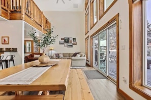 A spacious living area with a wooden dining table, a sofa, and a large window offering a view of the outdoors. - Lake House In Cascade w/ Hot Tub (Cascade)