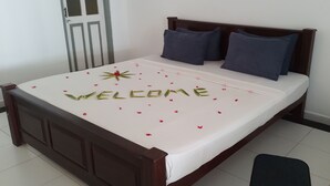 A neatly made bed with a white bedspread, two dark pillows, and a 'WELCOME' sign made of leaves and petals.