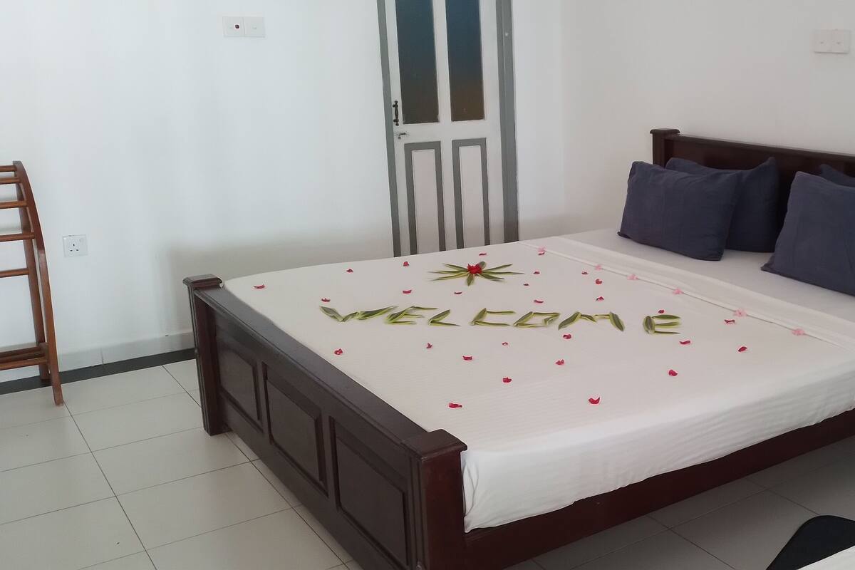 A neatly made bed with a 'WELCOME' sign and scattered rose petals, a wooden chair, and a door in a simple room with white walls and tiled flooring.