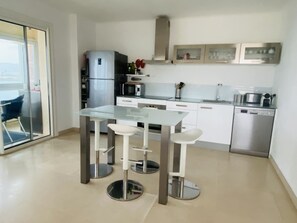 Une cuisine moderne avec une table à manger en verre, des appareils électroménagers en acier inoxydable et une porte-fenêtre coulissante donnant sur un balcon.