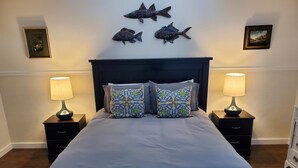 Un dormitorio con una cama, dos mesitas de noche con lámparas, un cabecero oscuro y tres piezas de arte mural con motivos de peces.