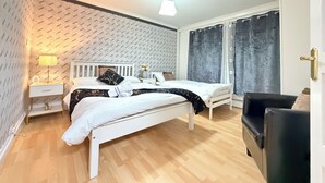 Un dormitori amb llit blanc, una nitòria, una làmpada, una finestra amb cortines i una porta.