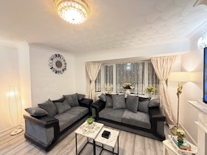 Uma sala de estar moderna com dois sofás cinzentos, uma pequena mesa de centro, uma peça decorativa na parede e uma grande janela com cortinas.