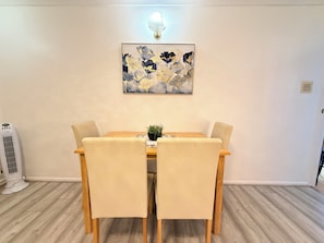 Uma sala de jantar com mesa de madeira e quatro cadeiras, um vaso com planta sobre a mesa e um candeeiro fixado na parede.
