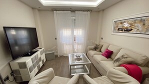 Ein modernes Wohnzimmer mit Flachbildfernseher, einem Glastisch, zwei weißen Sesseln, einem beige farbenen Sofa mit Zierkissen und einem großen abstrakten Gemälde an der Wand.