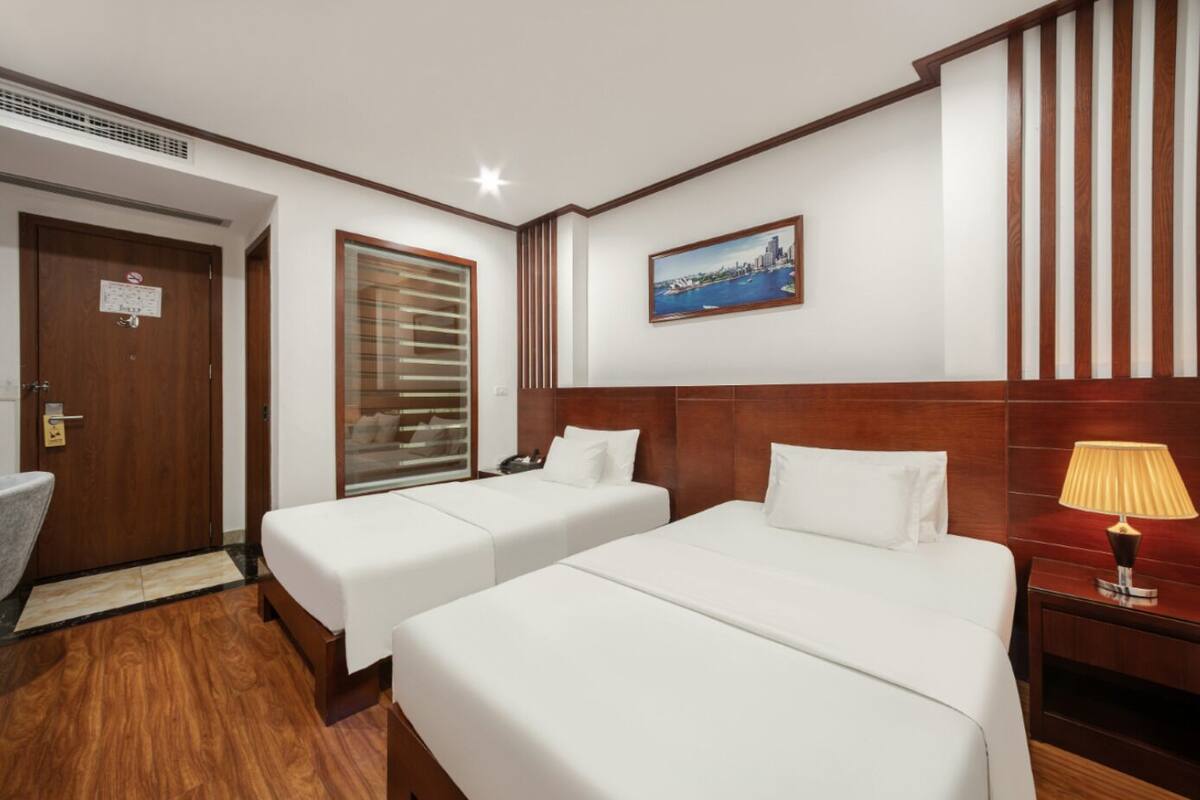 A hotel room with two single beds, a wooden headboard, a nightstand with a lamp, a window with blinds, and a painting on the wall.