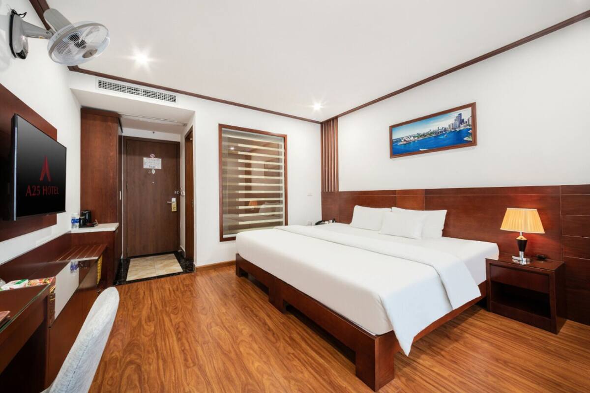 A hotel room with a bed, bedside lamp, TV, wooden floor, and a cityscape painting on the wall.