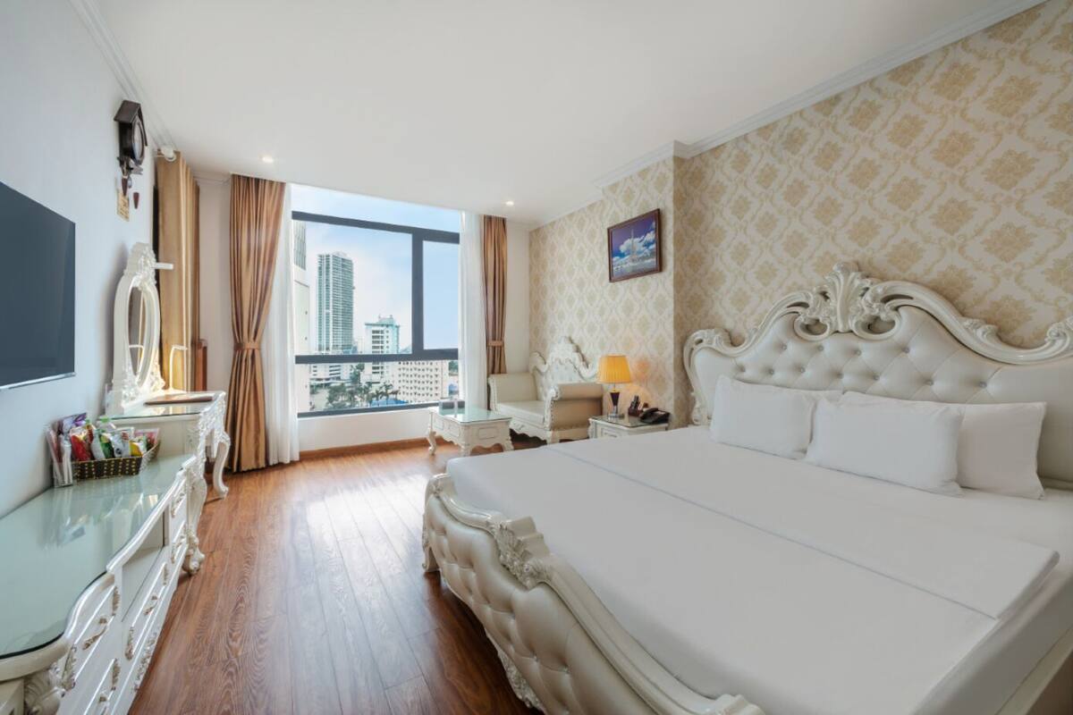 A hotel room with a large bed, a desk with a lamp, a chair, a TV, and a view of the cityscape.