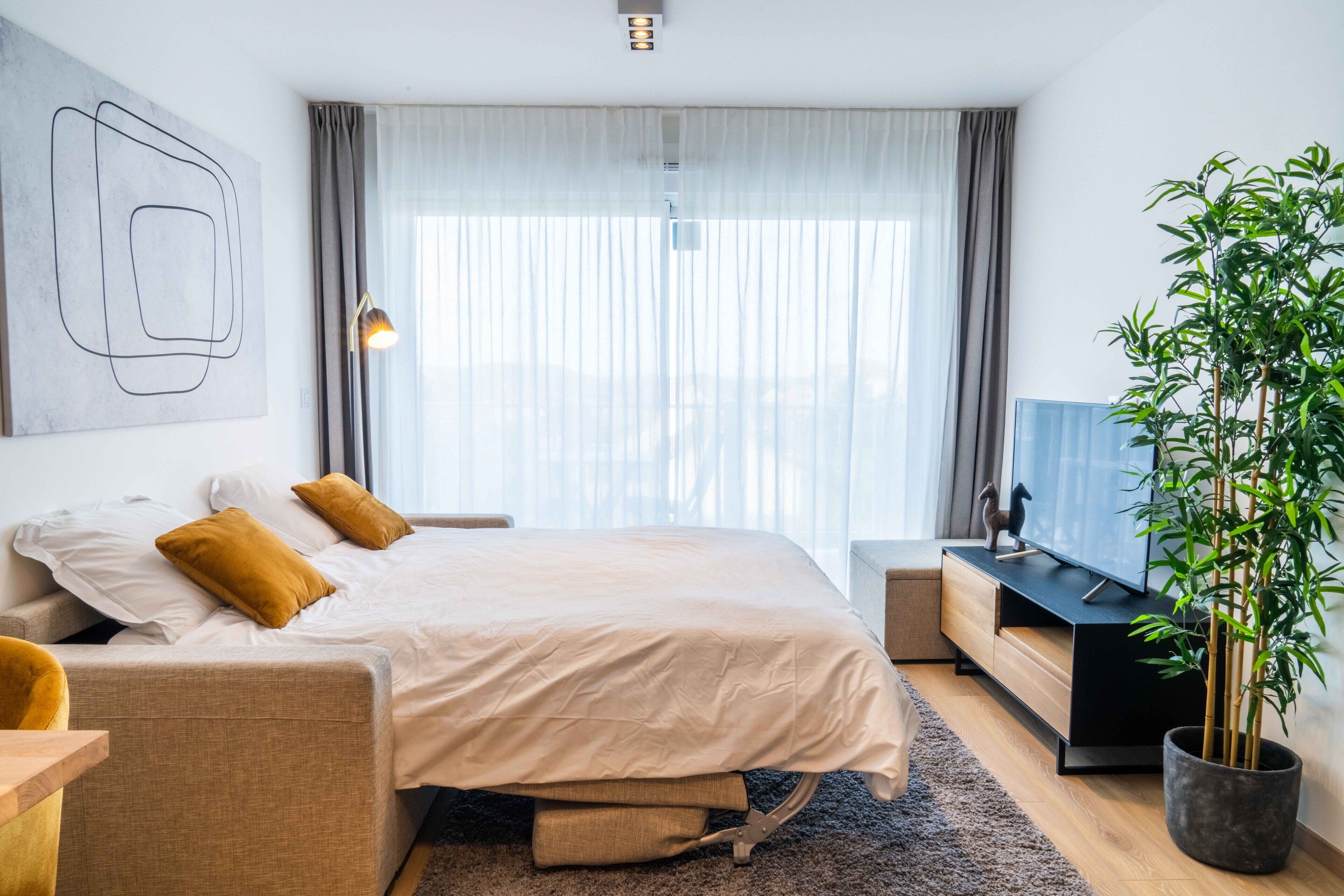 Moderni hotellihuone, jossa on sänky, työpöytä, televisio ja suuri ikkuna, jossa on verhot.