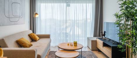 Una sala de estar moderna con un sofá, dos mesas de centro, un mueble para televisor y un ventanal con cortinas traslúcidas.