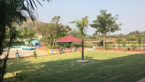 A well-maintained lawn with a red-roofed pavilion, a fenced area with a palm tree, and a building with a blue facade in the background. -  Royal Hills Resort (Mawal)