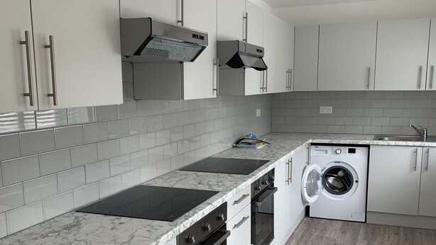 A modern kitchen with white cabinets, a black induction cooktop, a built-in oven, a washing machine, and a grey tiled backsplash. - Wagtail Way Kent (Orpington)
