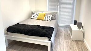 A bedroom with a bed, a bedside table, a lamp, and a window with blinds. - Wagtail Way Kent (Orpington)