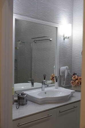 A bathroom with a large mirror, a sink, and a hairdryer. There is a shower visible in the background. - King David Residences (Tbilisi)