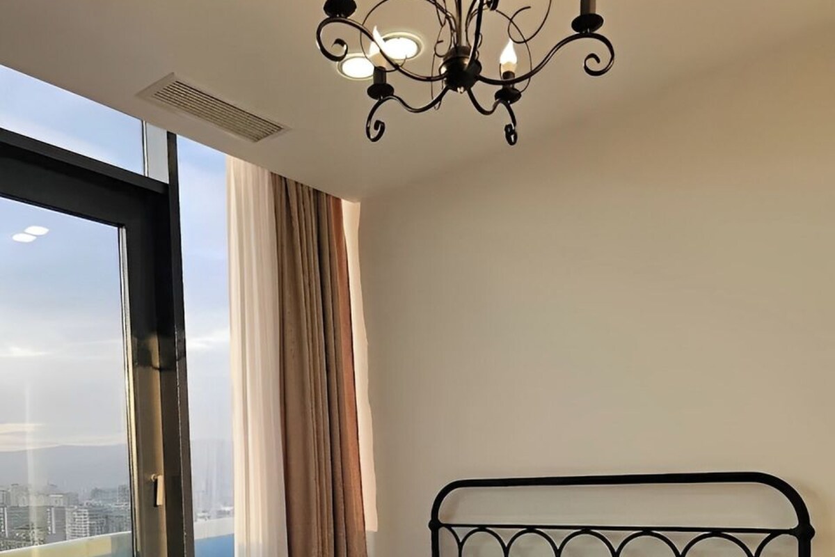 Quarto moderno com cama grande, cabeceira de metal preto, lustre e vista para a cidade através de uma janela ampla.