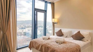A hotel room with a large window, a bed with a patterned bedspread, and a city view. - King David Residences (Tbilisi)