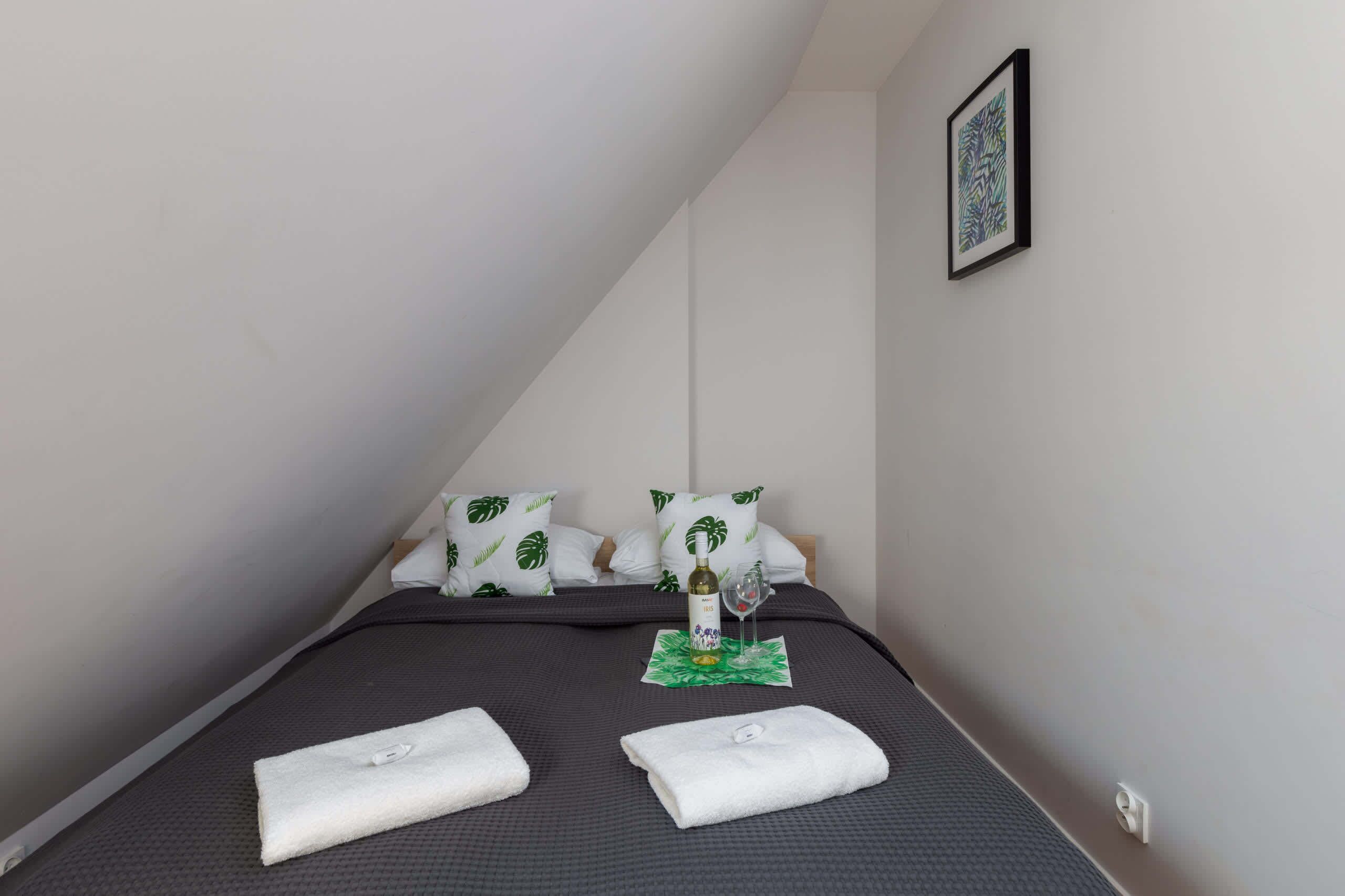 Un lit bien fait, avec une literie sombre, deux serviettes blanches, une bouteille et un verre posés sur un tapis vert.