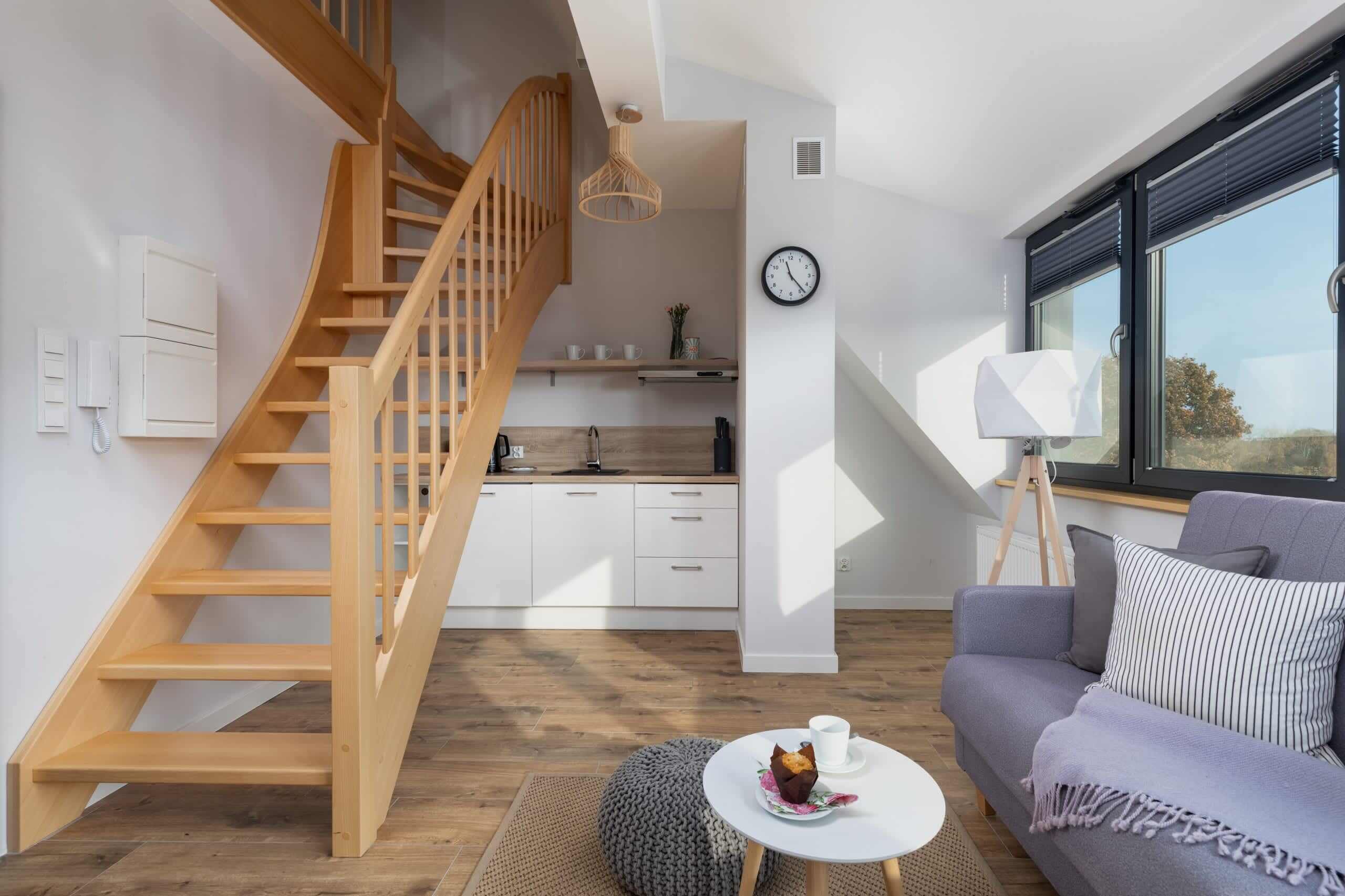 Un espacio habitable moderno con una escalera de madera, una zona de cocina, una mesa de comedor con una taza y un muffin, y un sofá con cojines.
