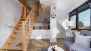 A modern living space with a wooden staircase, a kitchen area, a dining table with a cup and muffin, and a sofa with cushions.
