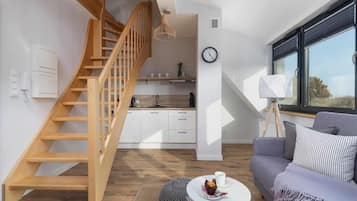Un espacio habitable moderno con una escalera de madera, una zona de cocina, una mesa de comedor con una taza y un muffin, y un sofá con cojines.