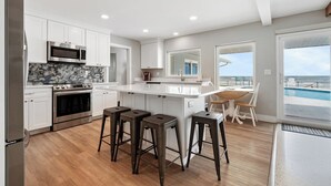 A modern kitchen with white cabinets, a central island with stools, a large window overlooking a pool, and a view of the ocean. - Ponce Inlet Beach House (Ponce Inlet)