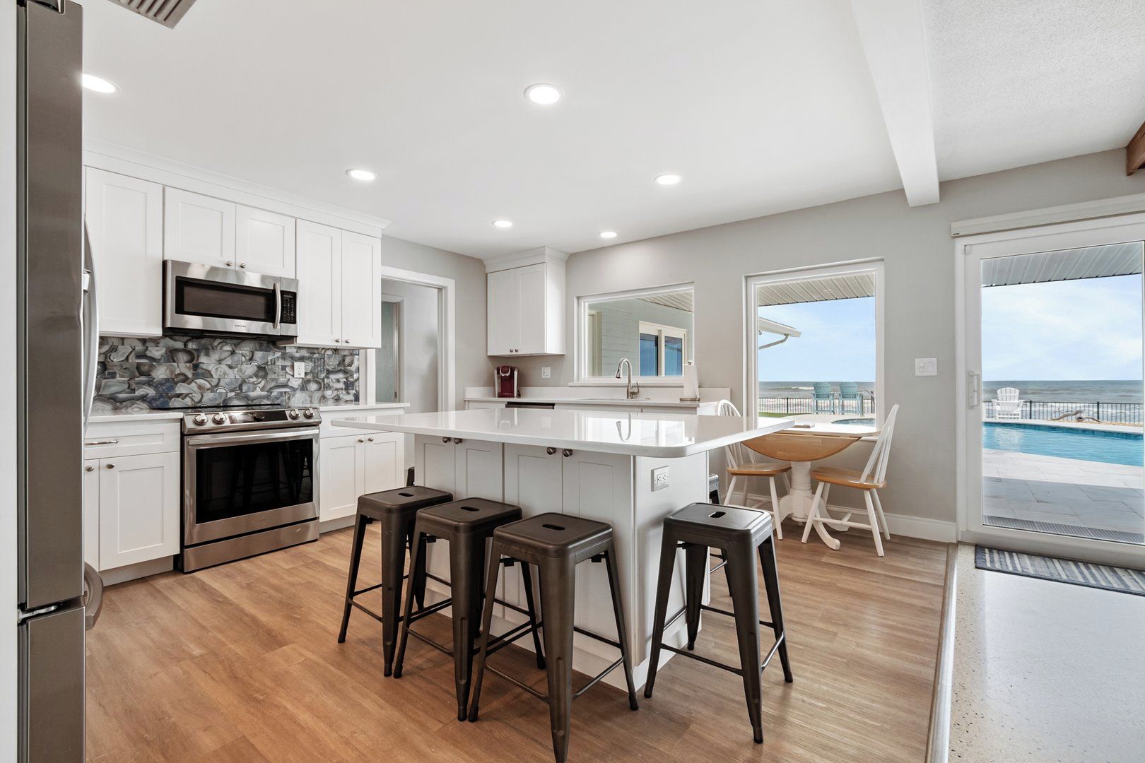 A modern kitchen with white cabinets, a central island with stools, a large window overlooking a pool, and a view of the ocean.