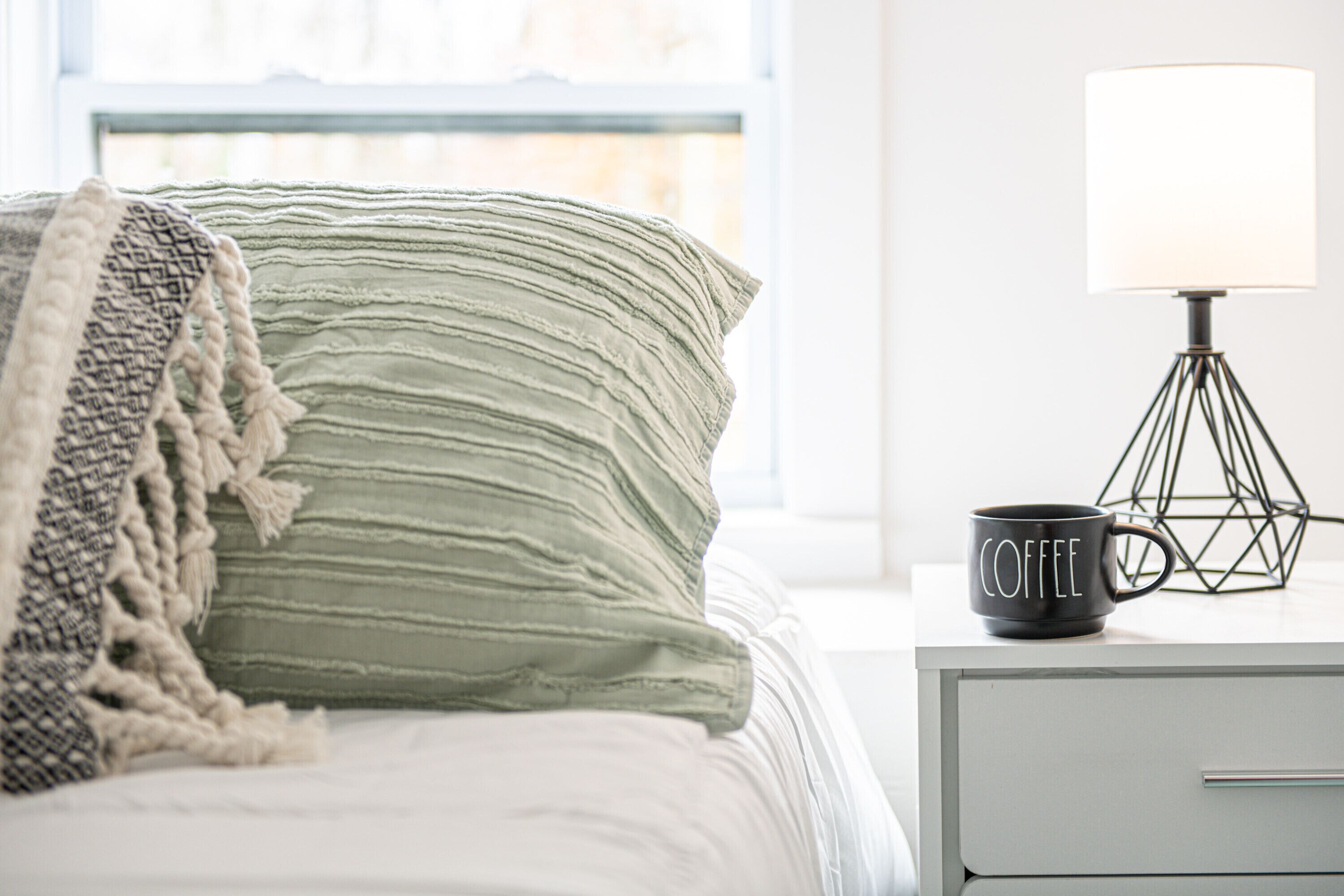 Un lit bien fait, avec un oreiller à rayures vertes et une couverture blanche, une tasse à café noire posée sur une table de chevet, et une lampe dont le pied est de forme géométrique.