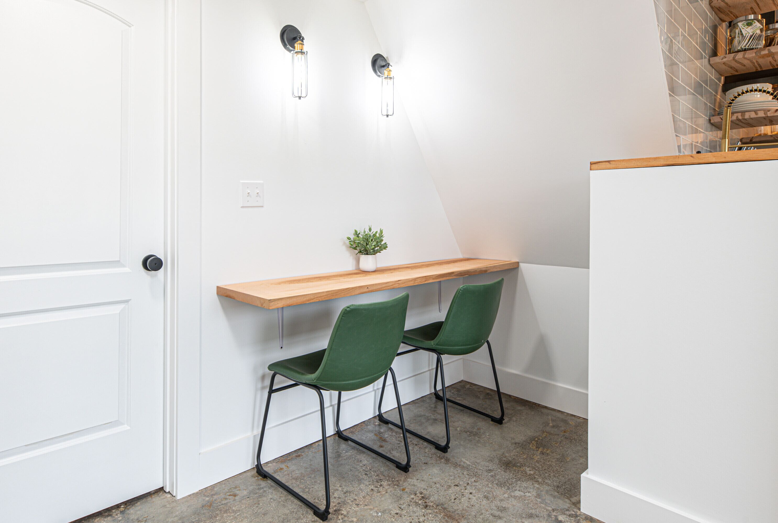 A small, clean room with a white door, a wooden desk, two green chairs, and two wall-mounted light fixtures.