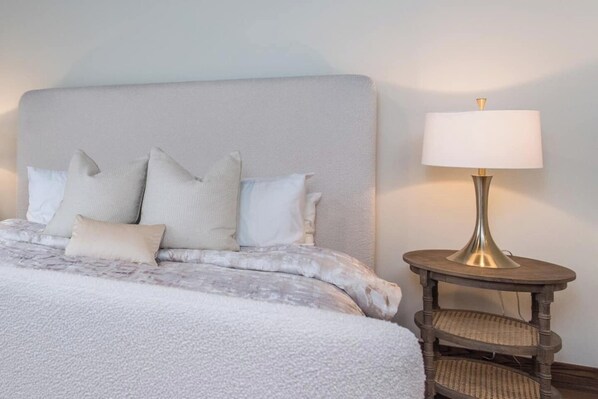A neatly made bed with a grey headboard and multiple pillows, a round wooden side table with a lamp, and a wicker chair.