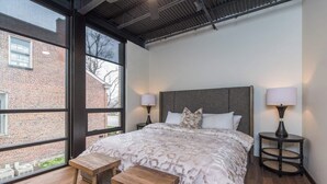 A bedroom with a large window, a bed with a patterned bedspread, two bedside tables with lamps, and a small wooden bench.