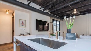 A modern kitchen with a white countertop, stainless steel sink, and a gold faucet. There is a black-framed window above the sink and a vase with white flowers on the countertop.