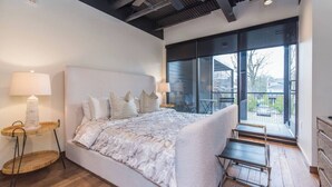 A modern bedroom with a large bed, a nightstand, a lamp, and a view of the outdoors through large windows.