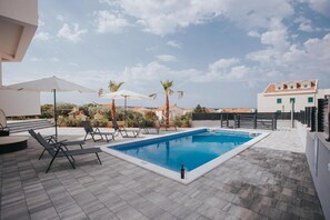Une piscine rectangulaire, agrémentée de chaises longues et de parasols, entourée d’une terrasse carrelée et de palmiers.