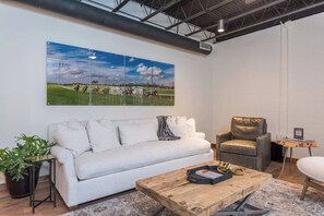 Ein modernes Wohnzimmer mit einem weißen Sofa, einem Ledersessel, einem Holztisch und einem großen gerahmten Bild von einem Pferderennen.