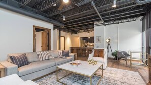 Ein modernes Wohnzimmer mit einer grauen Eckcouch, einem weißen Sessel und einem Couchtisch. Im Hintergrund ist eine Küche erkennbar.