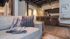 Ein modernes Wohnzimmer mit einem grauen Sofa, einem gestreiften Kissen und einem kleinen Teppich. Im Hintergrund ist eine Küchenzeile mit Holzbarhocker erkennbar.