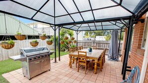 Un porche cubierto con una parrilla, mesa de madera, sillas, macetas colgantes y una manguera.