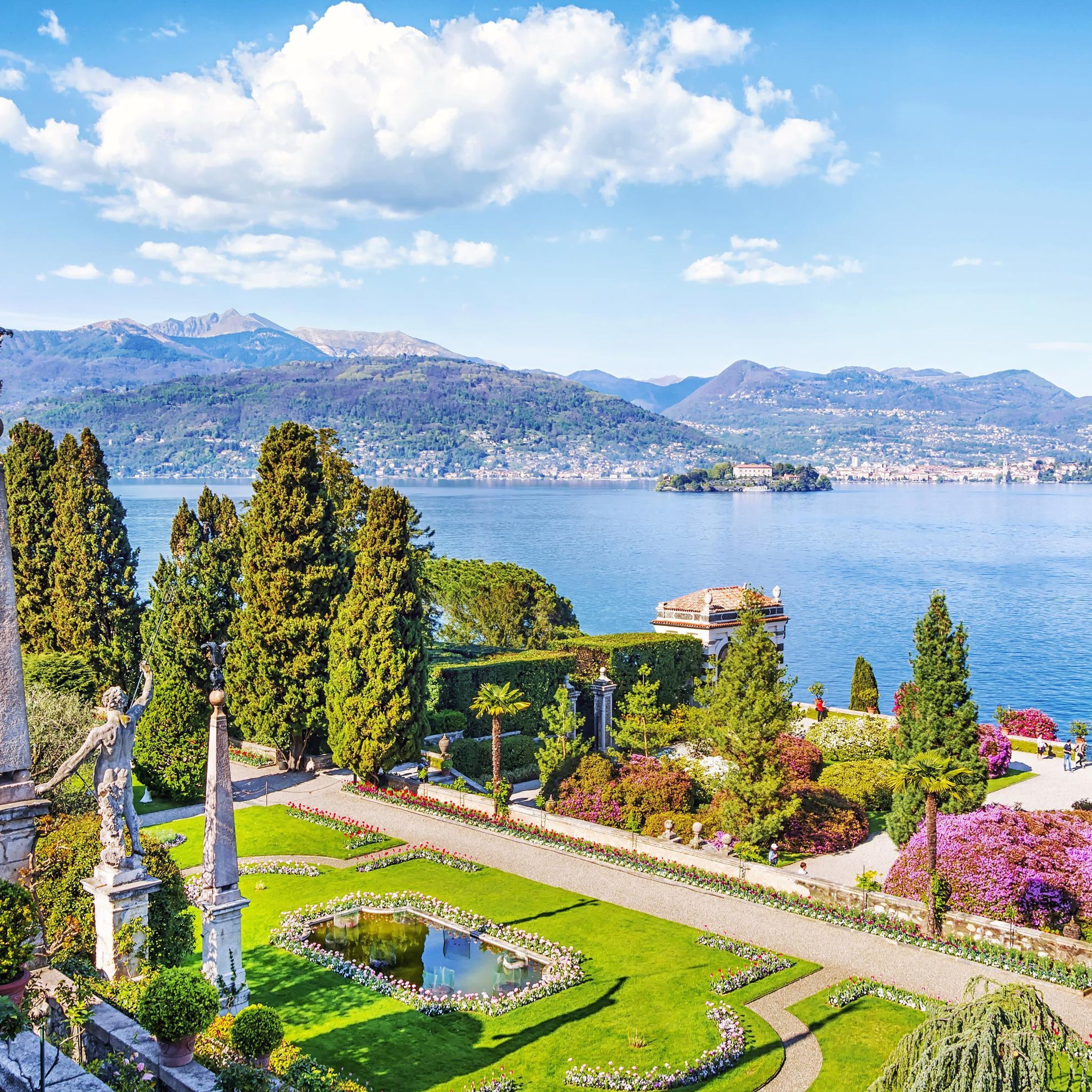 A landscaped garden with a central fountain, manicured lawns, and a view of a lake and distant mountains.