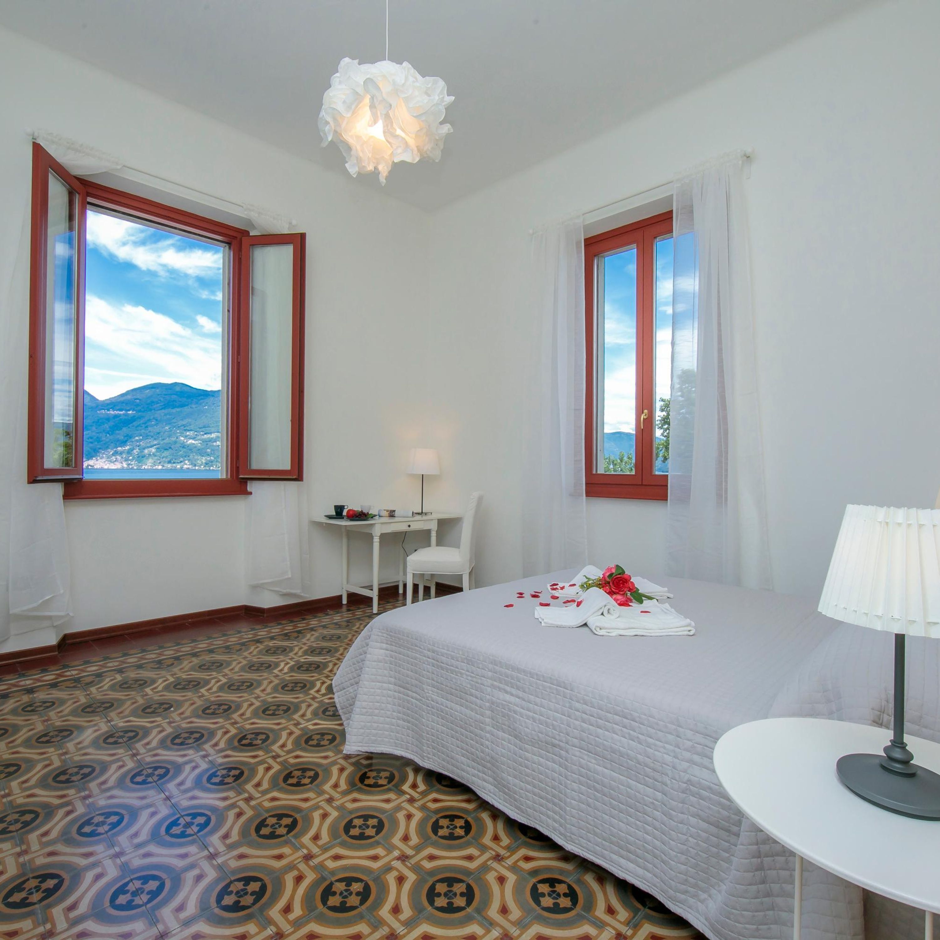 Habitación de hotel con cama, escritorio, silla, lámpara y ventanas grandes con cortinas.