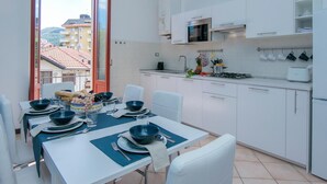 Una cucina moderna con mobili bianchi, un'area pranzo con tavolo apparecchiato per quattro persone e vista sugli edifici attraverso una porta aperta.