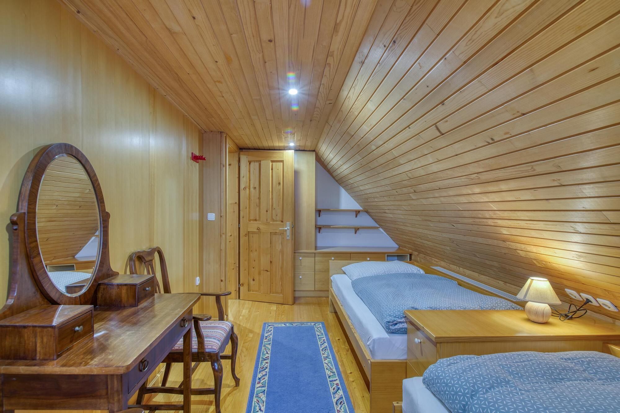Une pièce aux murs lambrissés, comprenant un bureau, une chaise, deux lits, un miroir et une petite table avec une lampe.