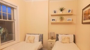 A bedroom with two beds, a window, a nightstand, and wall-mounted shelves with pictures and plants. - Clock Tower Flats Morpeth (Morpeth)