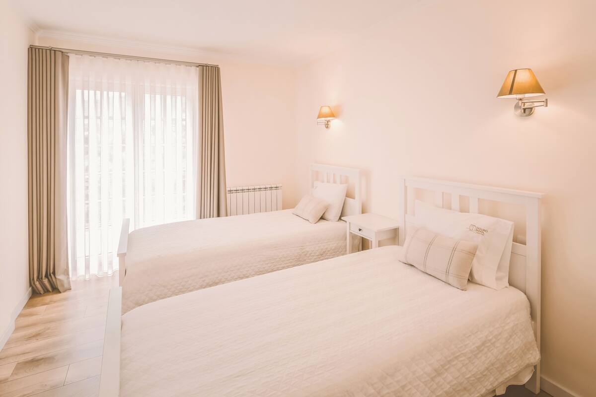 A hotel room with two beds, a window with curtains, a wall-mounted lamp, and a nightstand.