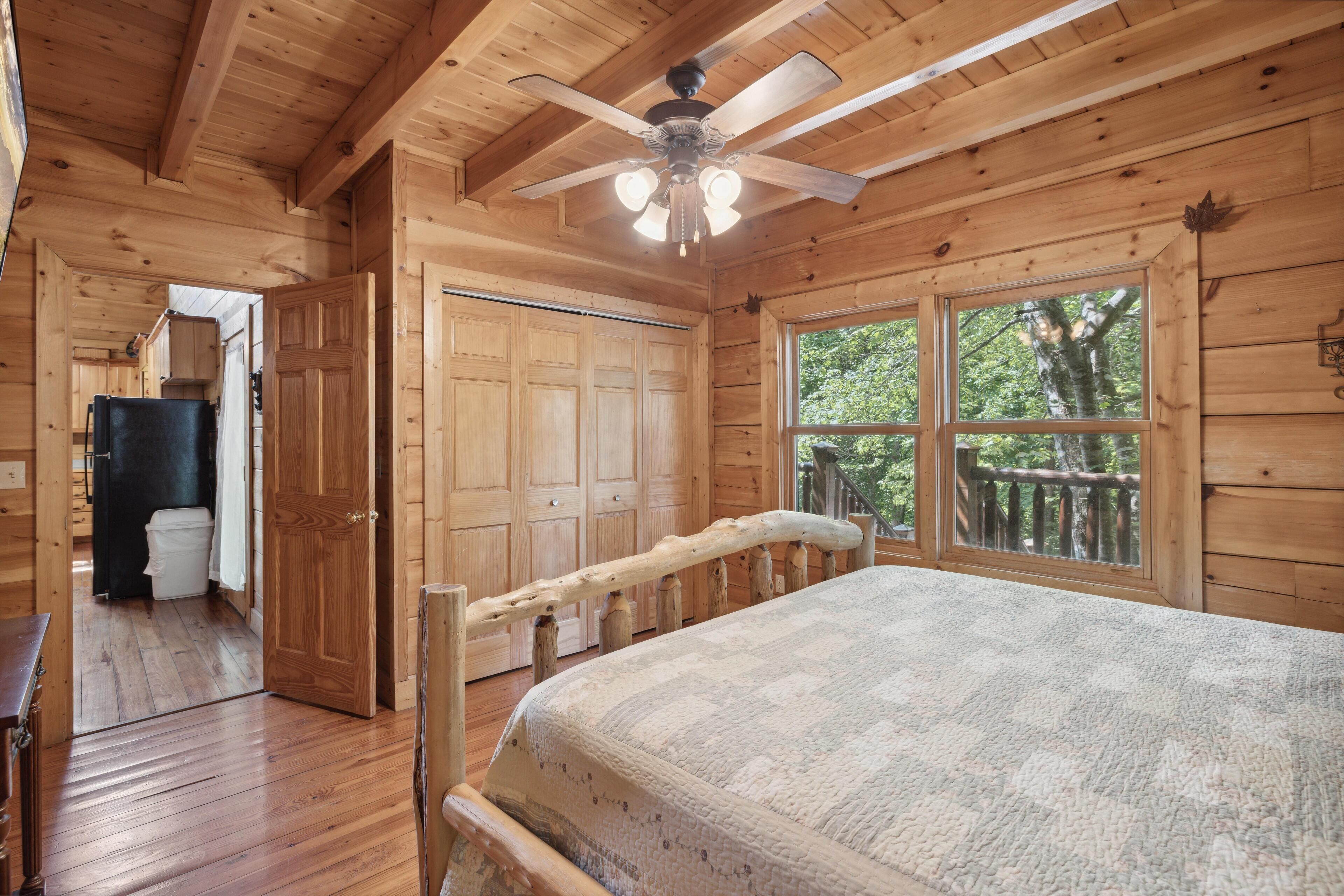 A wooden cabin bedroom with a bed, a ceiling fan, and a window with a view of trees.