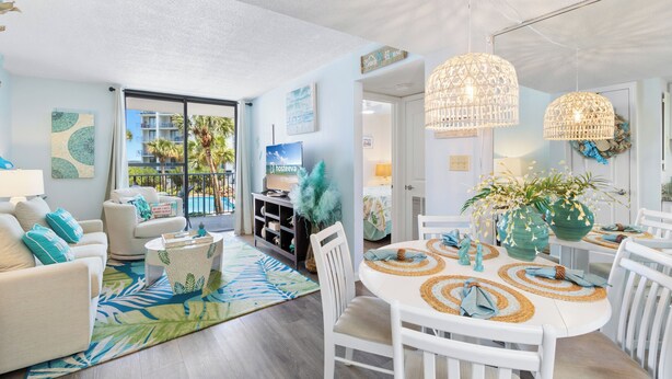 A modern living room with a dining area, featuring a large window with a view of palm trees and a pool. There are white furniture pieces, decorative plants, and a tropical-themed rug. - Salt and Light Surf & Racquet 204B (Gulf Shores)