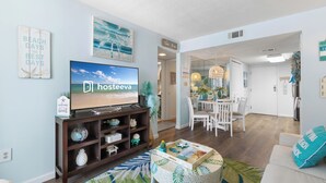 A modern living room with a flat-screen TV, a coffee table, and a dining area with a chandelier. - Salt and Light Surf & Racquet 204B (Gulf Shores)