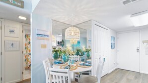 A dining area with a round table, white chairs, and a chandelier. - Salt and Light Surf & Racquet 204B (Gulf Shores)