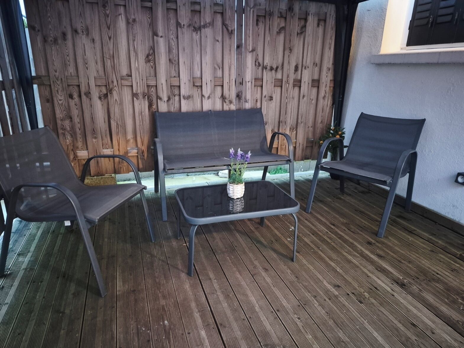 A patio with two metal chairs and a bench, a small table with a potted plant, and a wooden fence in the background.