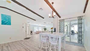 Un vaste espace de vie doté d’un plancher en bois, d’une table à manger avec des chaises blanches, d’un plafond avec des poutres apparentes et d’une vue sur l’extérieur grâce à une porte vitrée.